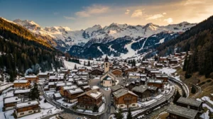 Vue aérienne d'un village alpin traditionnel au crépuscule avec chalets en bois disposés face aux sommets enneigés sous une lumière dorée